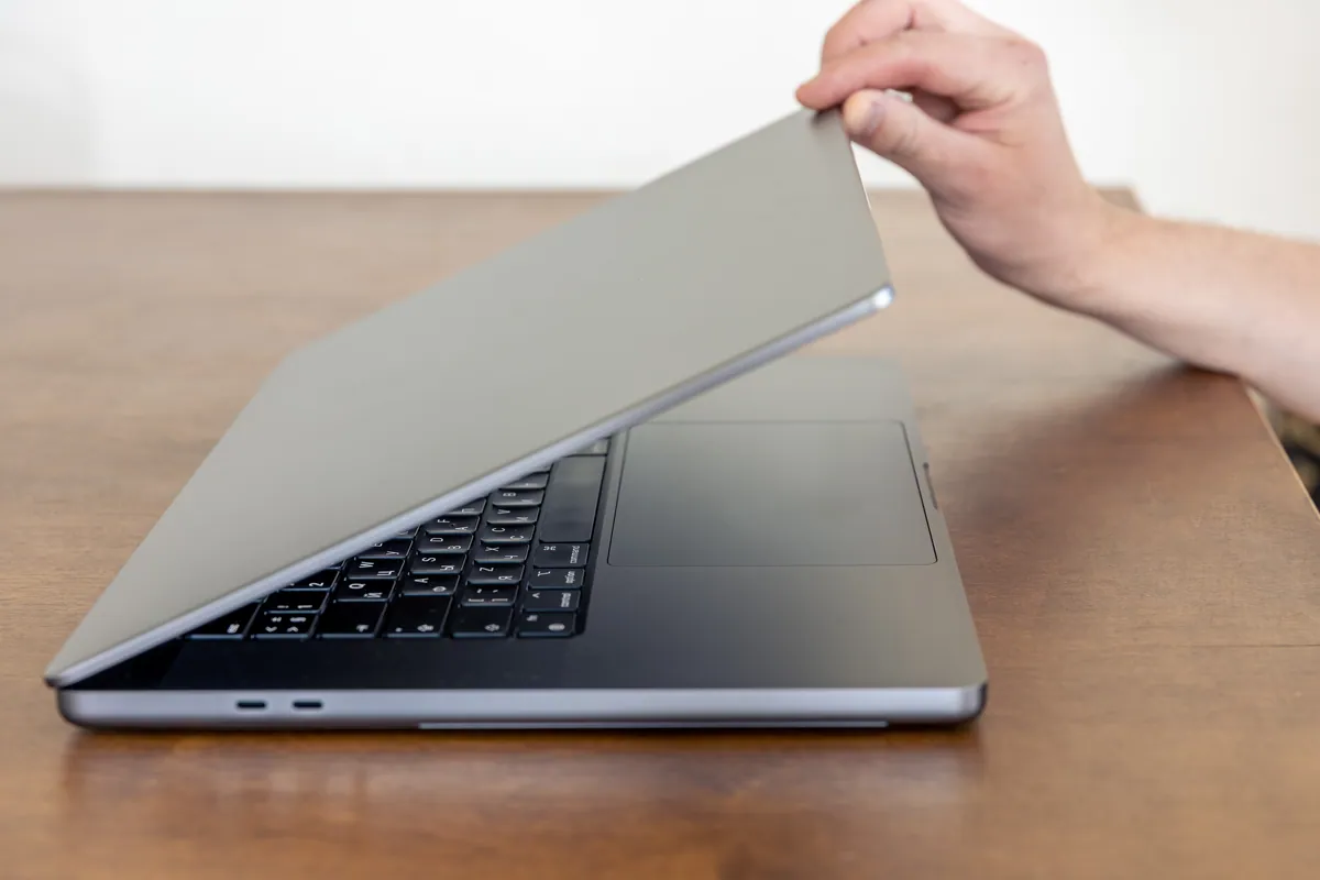 A hand lifting the lid of a silver laptop on a wooden desk